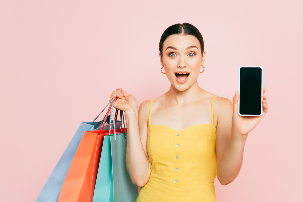 ショッピングバッグを持つショックを受けたブルネットの若い女性ピンクに隔離された空白の画面を持つスマートフォンを提示 - 写真・画像