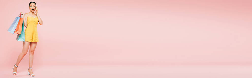 Ganzkörperansicht der schockierten brünetten jungen Frau mit Einkaufstaschen im Gespräch auf dem Smartphone auf rosa, Panoramaaufnahme - Foto, Bild