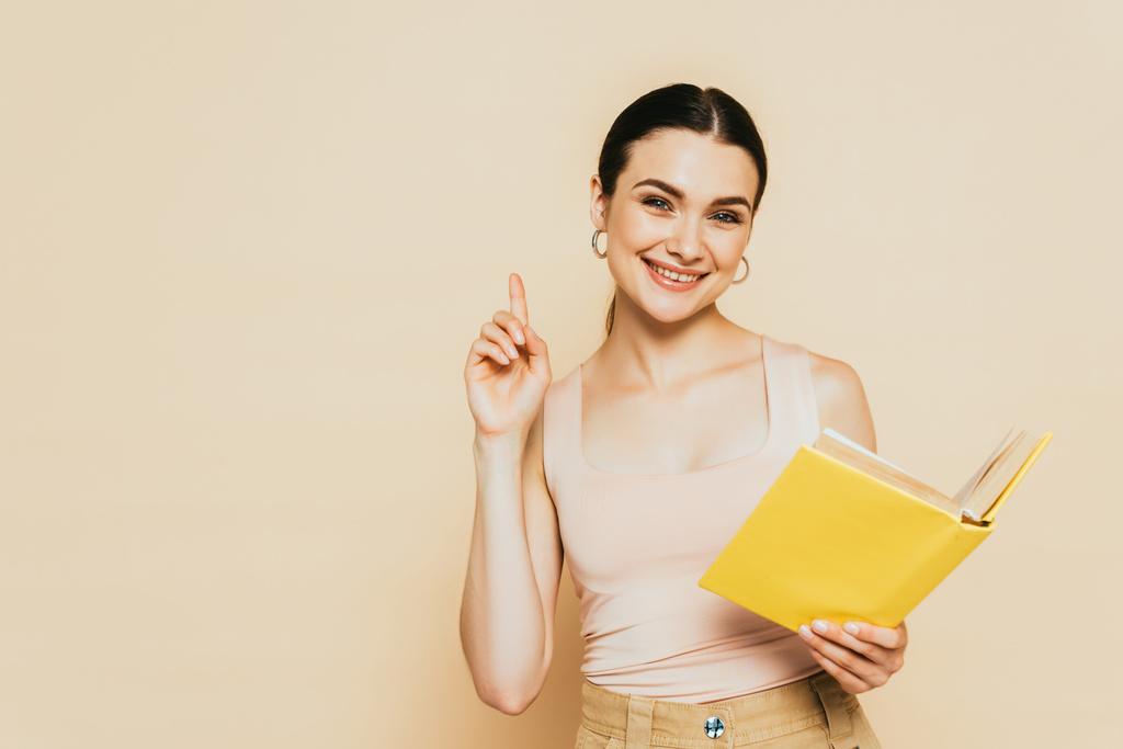 brünette junge Frau mit gelbem Buch zeigt Ideengeste isoliert auf beige - Foto, Bild