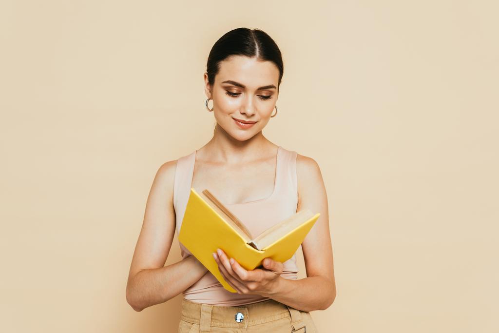 μελαχρινή νεαρή γυναίκα διαβάζει κίτρινο βιβλίο απομονωμένο σε μπεζ - Φωτογραφία, εικόνα
