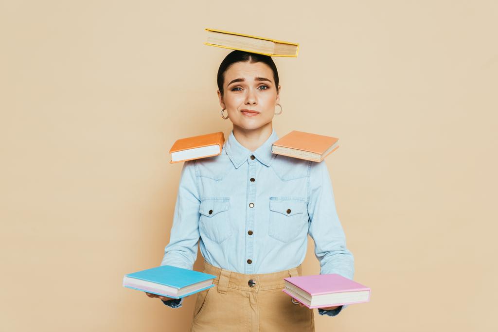 скептически настроенный студент в джинсовой рубашке с книгами по телу, изолированными на бежевом - Фото, изображение