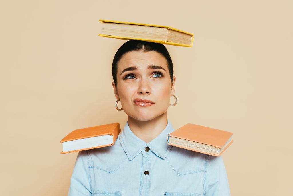 sceptische student in denim shirt met boeken op hoofd en schouders geïsoleerd op beige - Foto, afbeelding