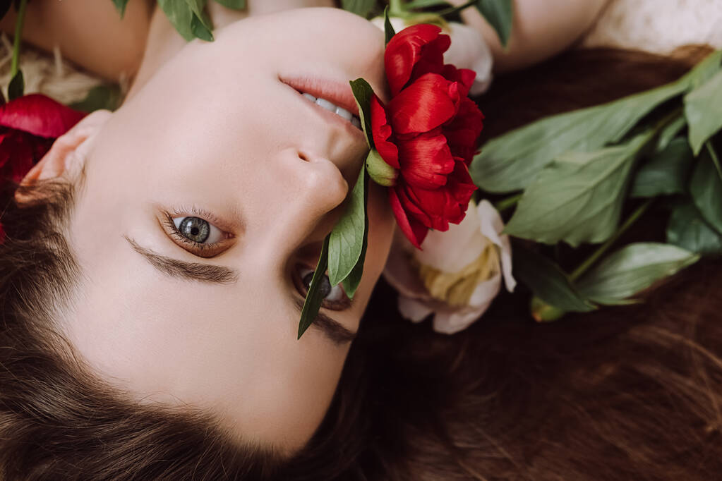 Seitenansicht Schönheit Porträt der attraktiven jungen brünetten Frau mit Blumen rote Pfingstrosen und grüne Blätter, Blick in die Kamera. Nahaufnahme Gesicht der schönen netten Mädchen mit gesunder, sauberer Haut - Foto, Bild