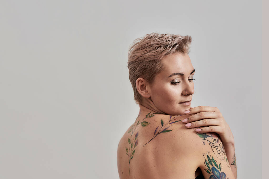 Ternura. Retrato de beleza de uma jovem atraente mulher tatuada semi-nua com a pele perfeita olhando para longe isolado sobre fundo cinza
 - Foto, Imagem