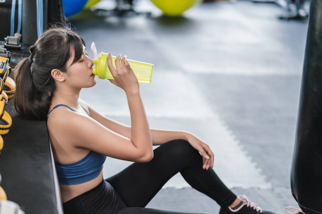 Молодая женщина после успешной тренировки в фитнес-зале держит бутылку белковой встряски и пьет, здоровый образ жизни бодибилдинг сидя на полу, концепция спортсмена наращивает мышцы. - Фото, изображение