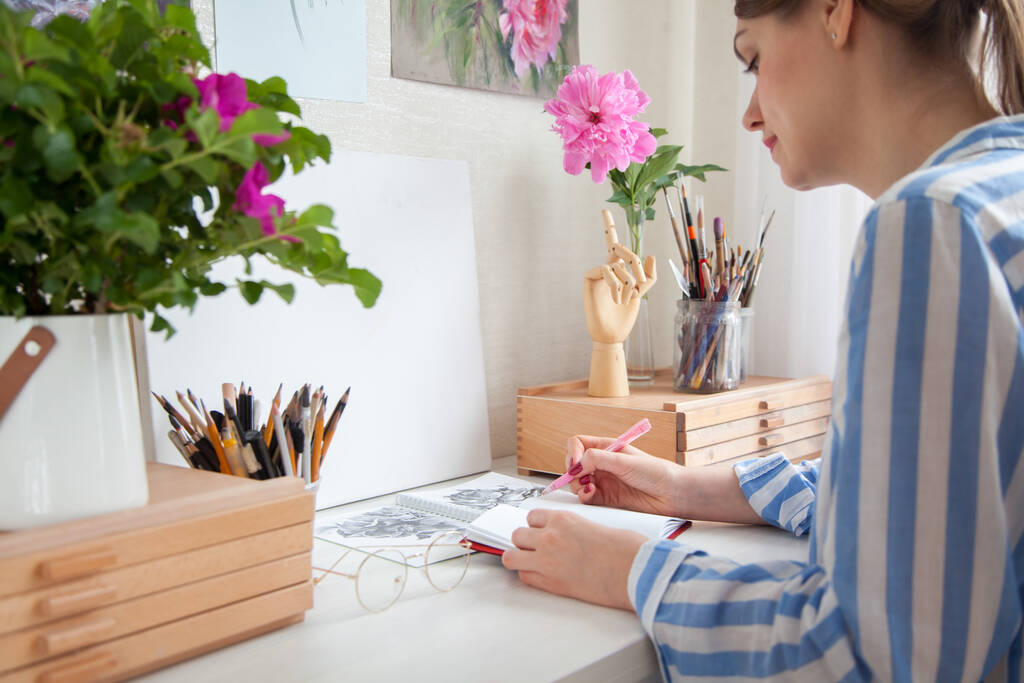 Вид збоку молодої кавказької дівчини художник заповнює щоденний планувальник пише список завдань у блокноті, сидячи за робочим столом з висувними ящиками з матеріалами для творчості. Творчість і концепція хобі
 - Фото, зображення