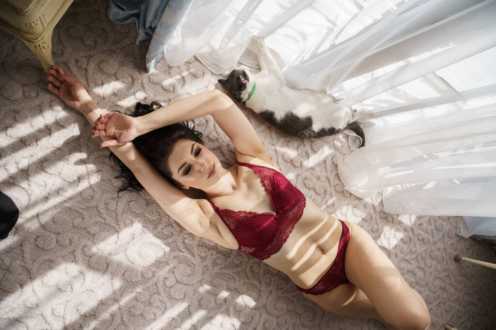 Sexy bruneta s kudrnatými vlasy hrát s kočkou v apartmánech interiéru. Portrét mladého modelu v červeném spodním prádle  - Fotografie, Obrázek