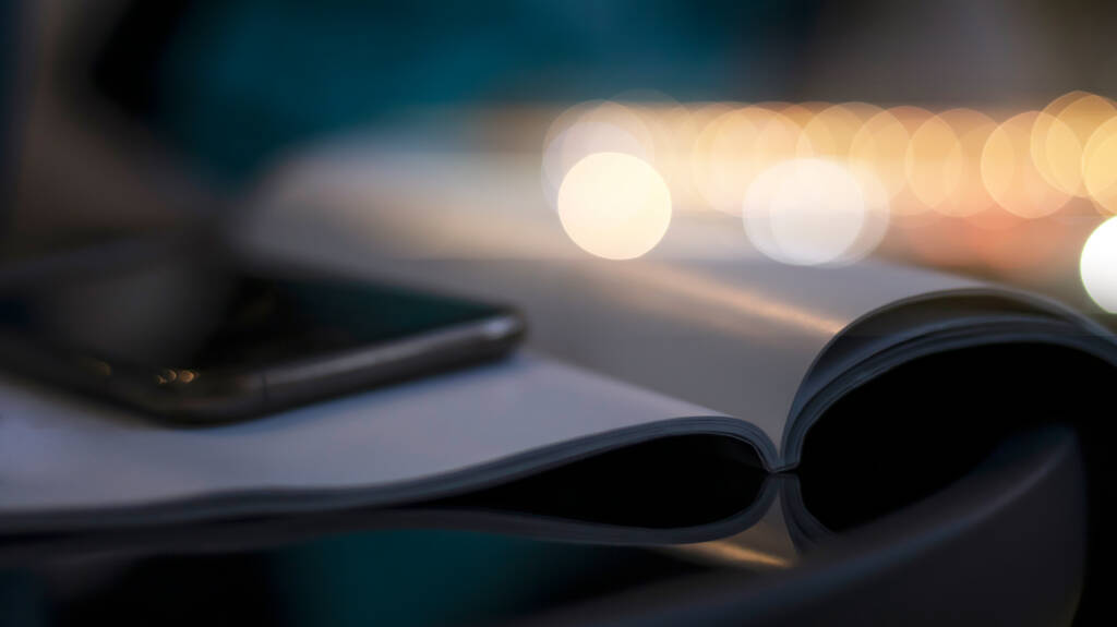 Open boek en telefoon op een zwarte glazen tafel. Wazige boke achtergrond, close-up. donkere avond achtergrond met zonsondergang hoogtepunten. - Foto, afbeelding