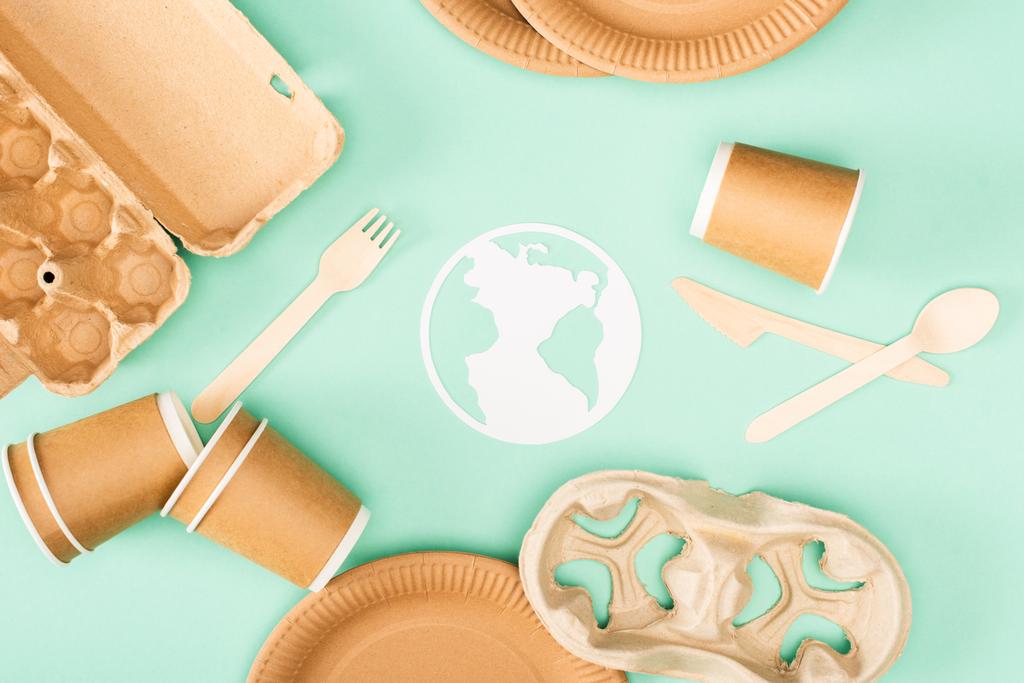Вид сверху бумажного знака земли рядом с одноразовой посудой и деревянной посудой на зеленом фоне, концепция экологии - Фото, изображение