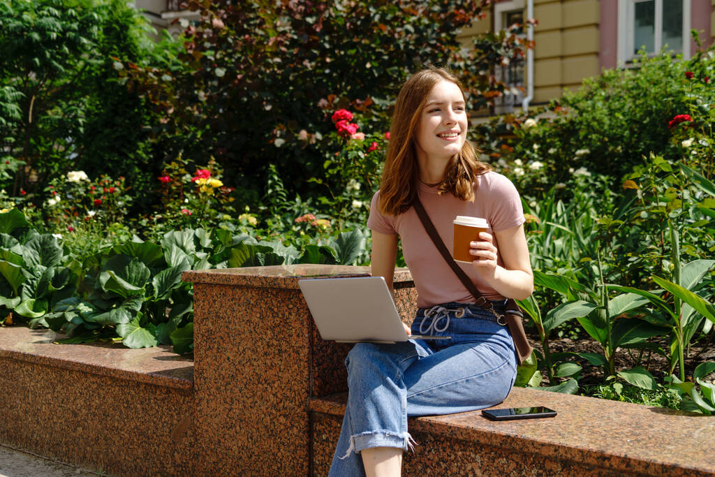 Όμορφη νεαρή γυναίκα τουρίστρια με takeout καφέ στο κέντρο της πόλης χρησιμοποιώντας φορητό υπολογιστή - Φωτογραφία, εικόνα