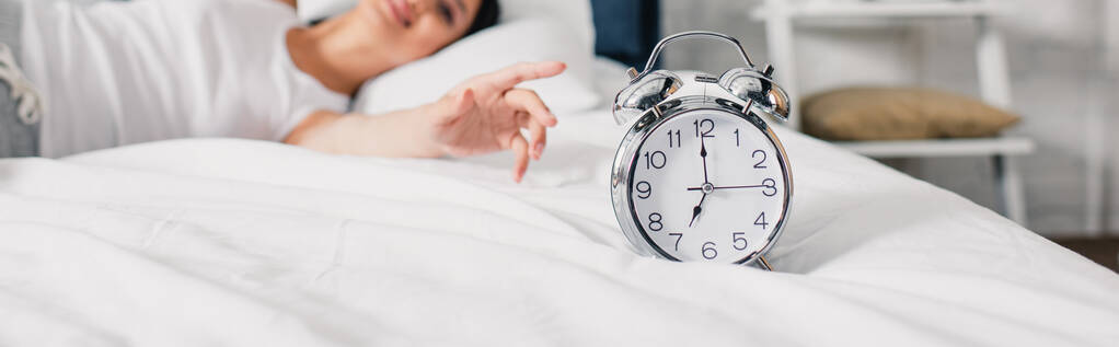 Vaaka sato herätyskello ja hymyilevä aasialainen nainen vetämällä käsi sängyllä aamulla  - Valokuva, kuva
