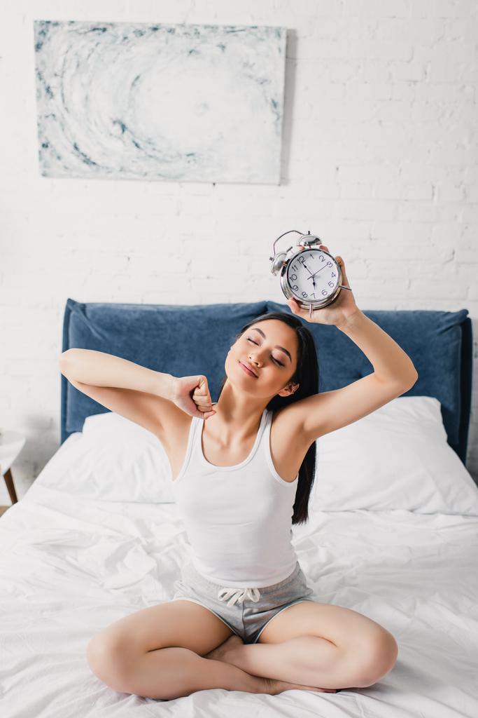 jonge aziatische vrouw met wekker en stretching op bed in de ochtend  - Foto, afbeelding