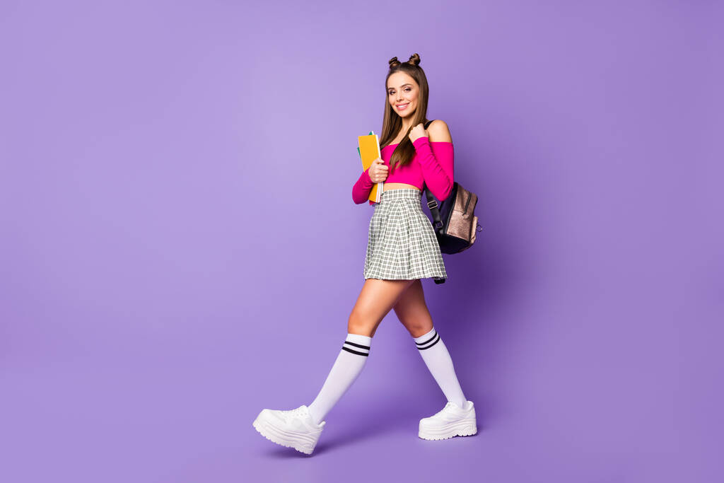 Полноразмерный вид на красивую симпатичную симпатичную симпатичную женственную жизнерадостную девочку, идущую в школу с книгой, изолированной на ярком ярком светящемся лилово-фиолетовом фоне - Фото, изображение