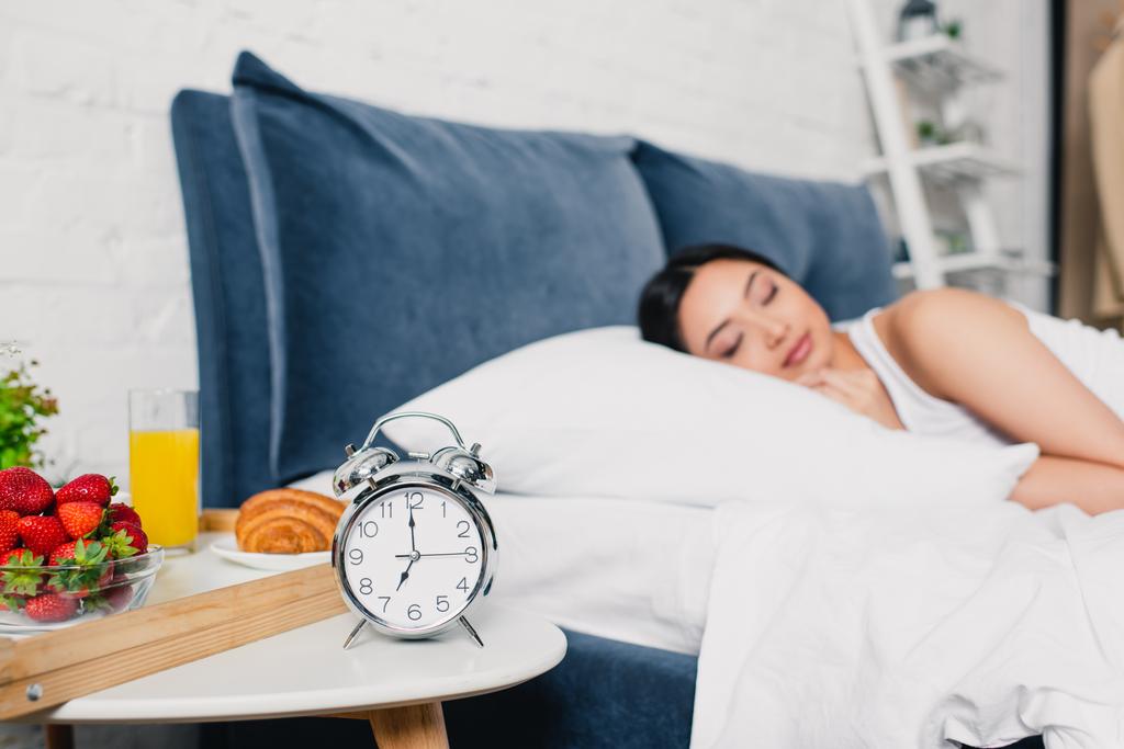 Вибірковий фокус будильника і сніданок на тумбочці і азіатська жінка спить на ліжку
  - Фото, зображення
