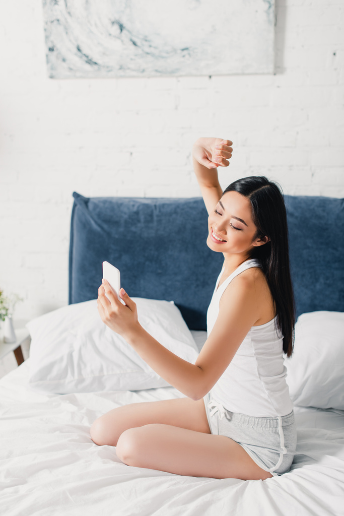 Sonriendo mujer asiática usando teléfono inteligente en la cama durante la mañana   - Foto, Imagen