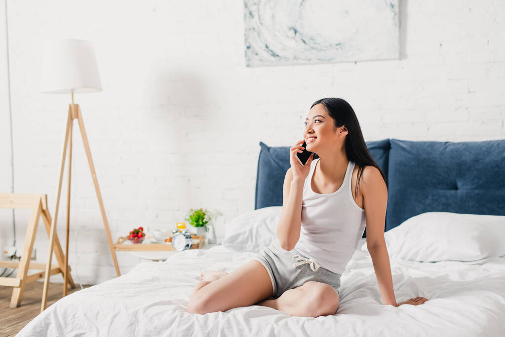 Atractiva chica asiática sonriendo y hablando en el teléfono inteligente en la cama por la mañana  - Foto, imagen