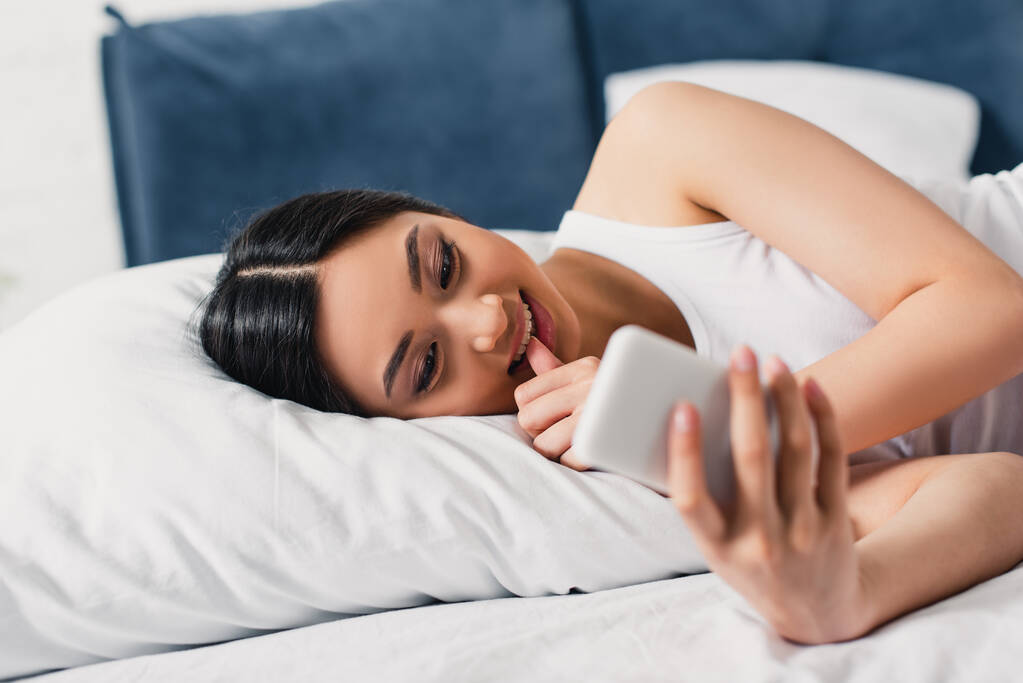 Selektywne skupienie uśmiechniętej azjatki z palcem w pobliżu ust przy użyciu smartfona na łóżku  - Zdjęcie, obraz
