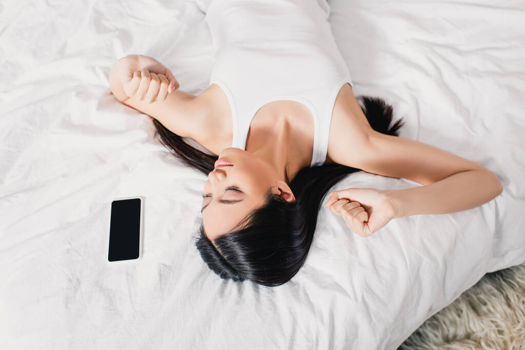 Top-Ansicht der asiatischen Frau Stretching in der Nähe von Smartphone auf dem Bett  - Foto, Bild