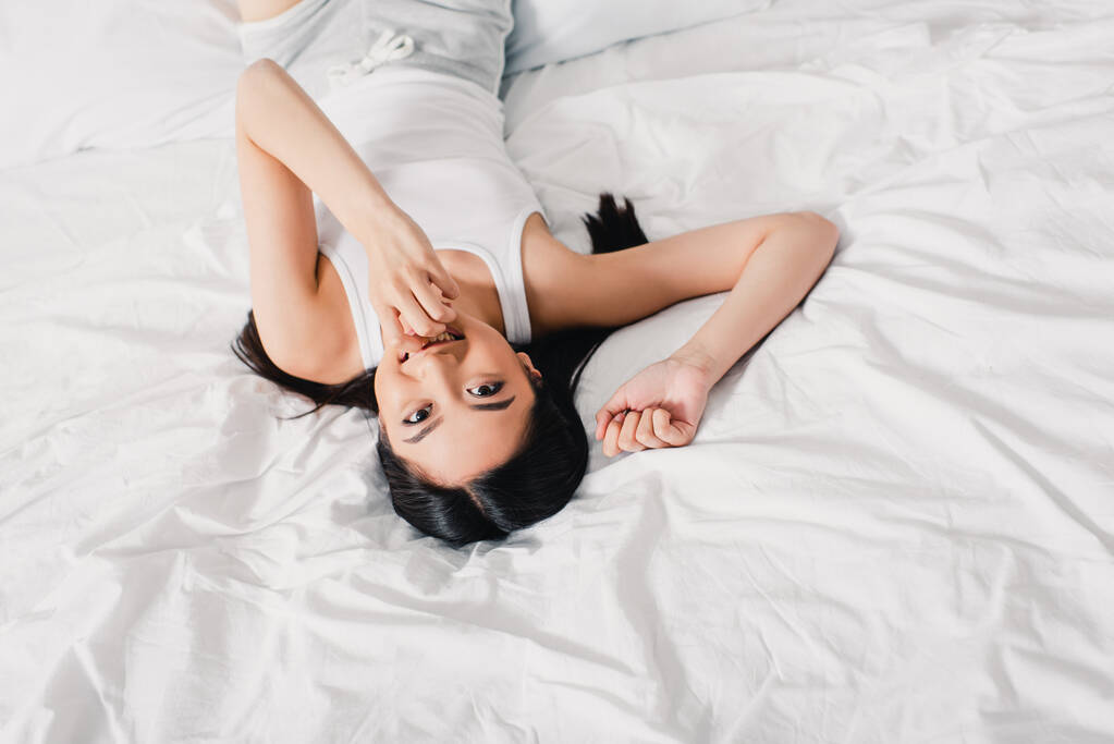 Duży kąt widzenia azjatyckiej dziewczyny z ręką w pobliżu ust uśmiecha się do kamery na łóżku  - Zdjęcie, obraz
