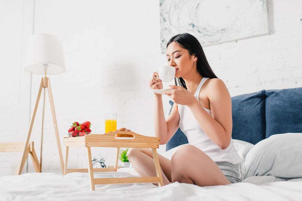 Ασιάτισσα γυναίκα πίνοντας καφέ κοντά στο δίσκο πρωινού στο κρεβάτι  - Φωτογραφία, εικόνα