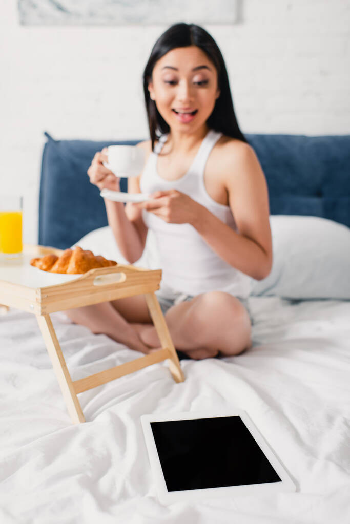 Foco seletivo de mulher asiática positiva segurando xícara de café e olhando para tablet digital na cama
  - Foto, Imagem