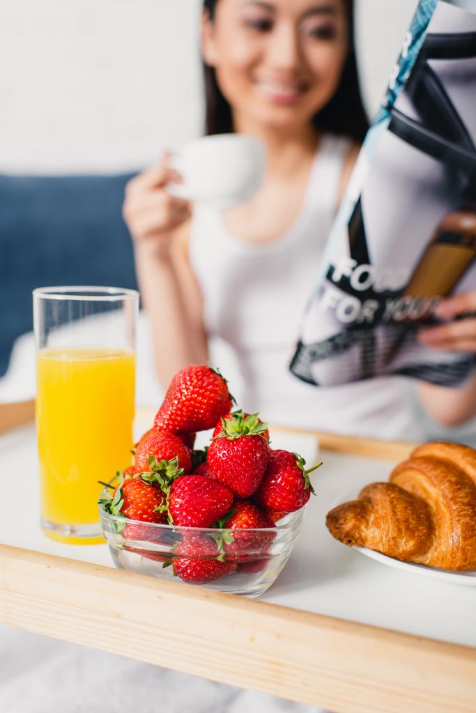 Selektywne skupienie śniadania na tacy i kobiecie trzymającej filiżankę kawy i czasopismo na łóżku  - Zdjęcie, obraz