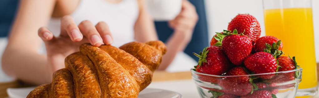 Panoramic crop of woman taking croissant near fresh strawberries and glass of orange juice  - Photo, Image