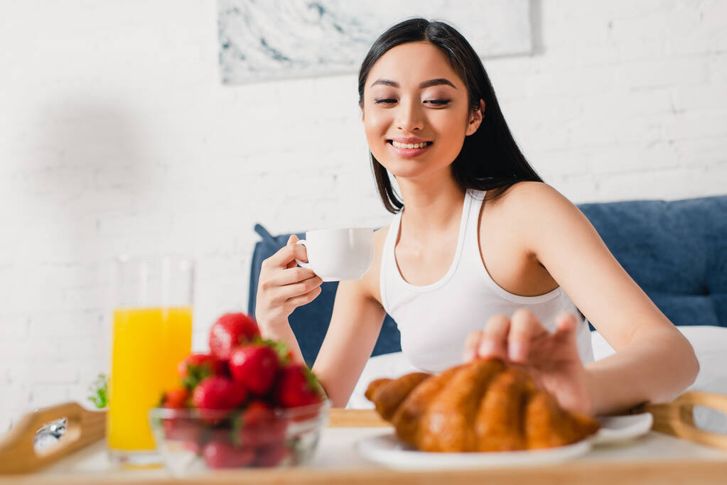 Вибірковий фокус усміхненої азіатської дівчини, яка приймає круасан під час сніданку на ліжку
  - Фото, зображення