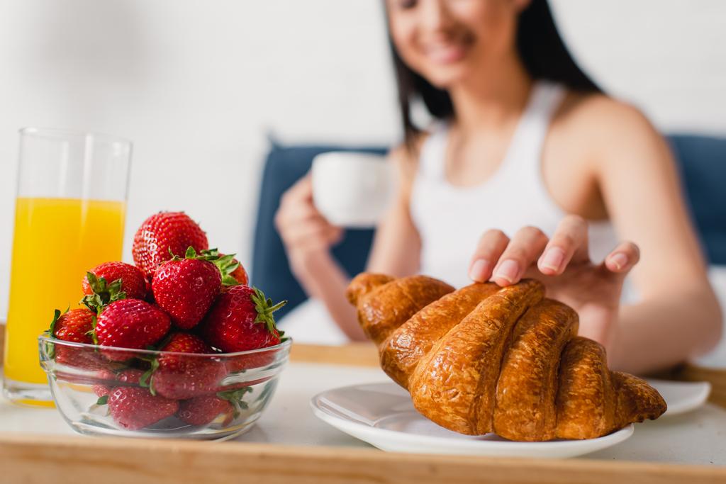 Selective focus of woman taking croissant near strawberries and glass of orange juice on breakfast tray  - Photo, Image