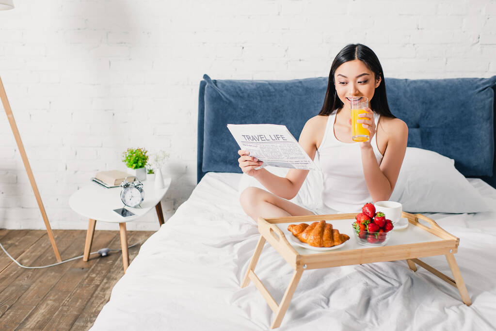 Valikoiva painopiste hymyilevä aasialainen tyttö käsittelyssä sanomalehti aamiaisen aikana appelsiinimehua ja croissant makuuhuoneessa - Valokuva, kuva