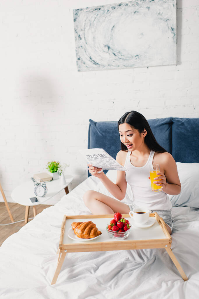 Joyeux asiatique fille lecture journal près du petit déjeuner sur plateau de petit déjeuner sur lit à la maison  - Photo, image