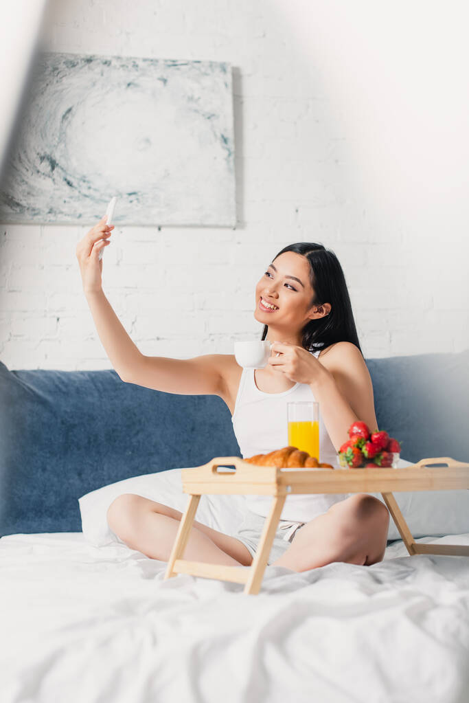 Selectieve focus van lachende Aziatische vrouw nemen selfie en houden koffie kopje in de buurt van het ontbijt op bed  - Foto, afbeelding