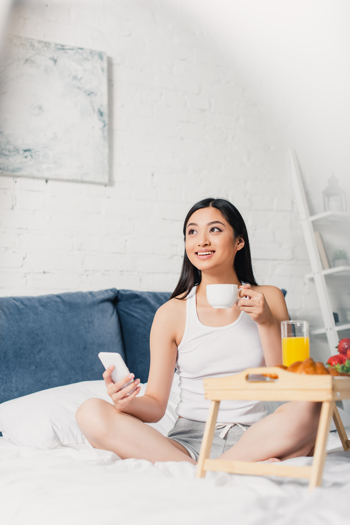 Selectieve focus van lachende Aziatische vrouw met kopje koffie en smartphone in de buurt van het ontbijt op bed  - Foto, afbeelding