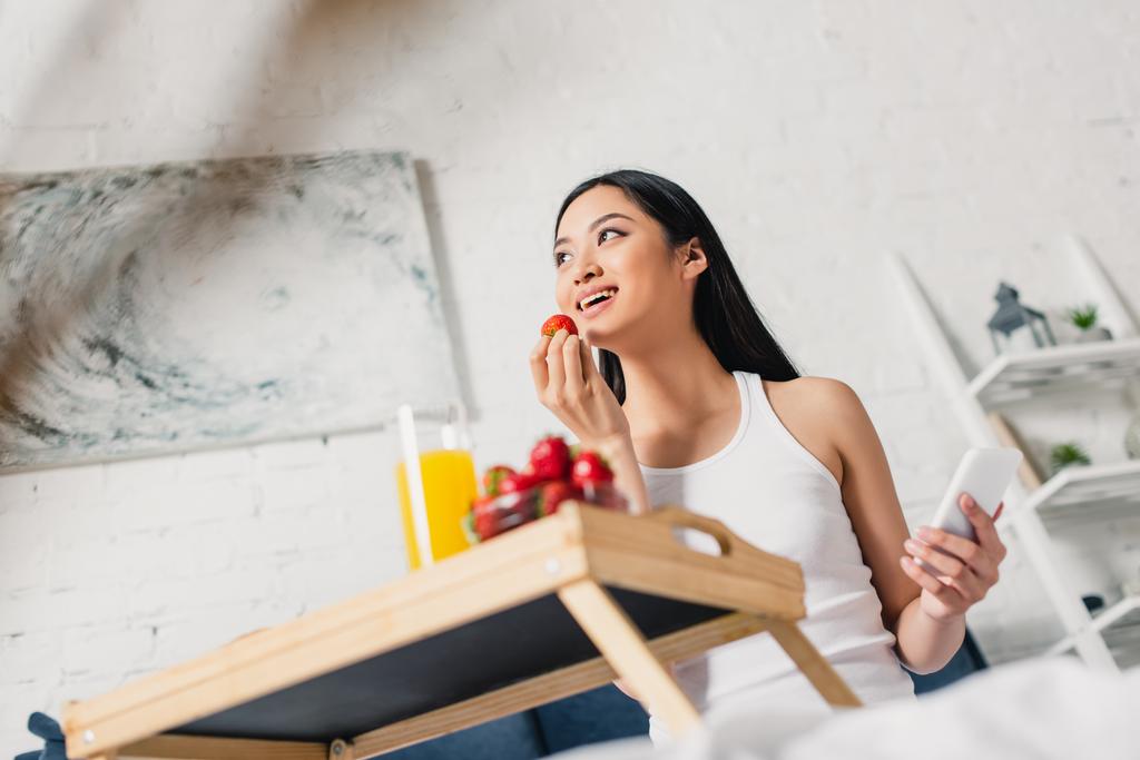 Selektywne skupienie się na uśmiechniętej azjatyckiej dziewczynie trzymającej truskawki i smartfona podczas śniadania w łóżku  - Zdjęcie, obraz