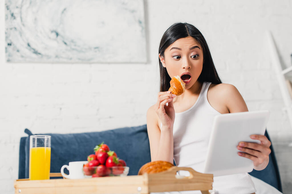 Selektiver Fokus einer überraschten Asiatin, die ein Stück Croissant und ein digitales Tablet in der Nähe des Frühstücks im Bett hält  - Foto, Bild