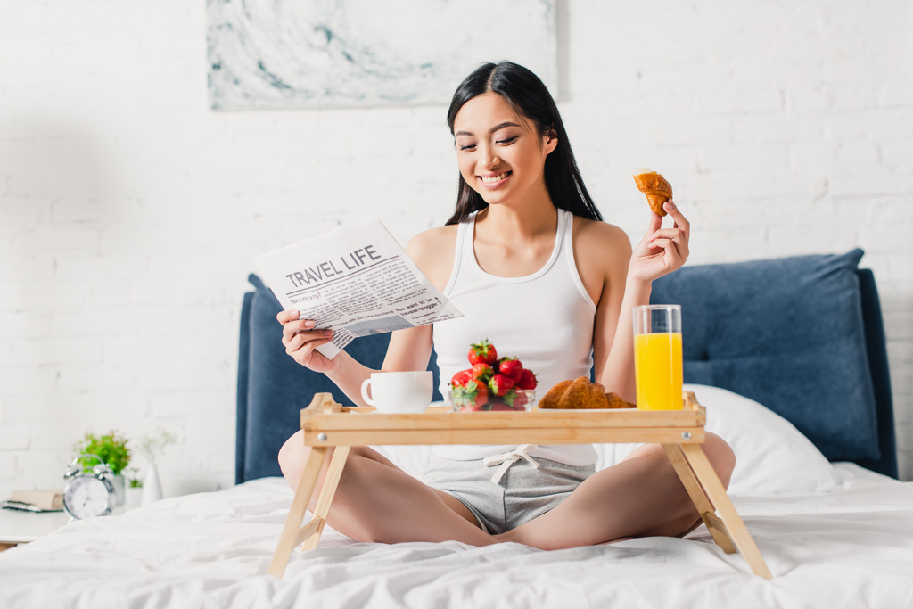 Foco seletivo de mulher asiática alegre ler jornal e segurando pedaço de croissant na cama
  - Foto, Imagem