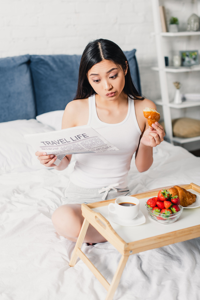 Foco seletivo de mulher asiática lendo jornal e tomando café da manhã na cama em casa
  - Foto, Imagem