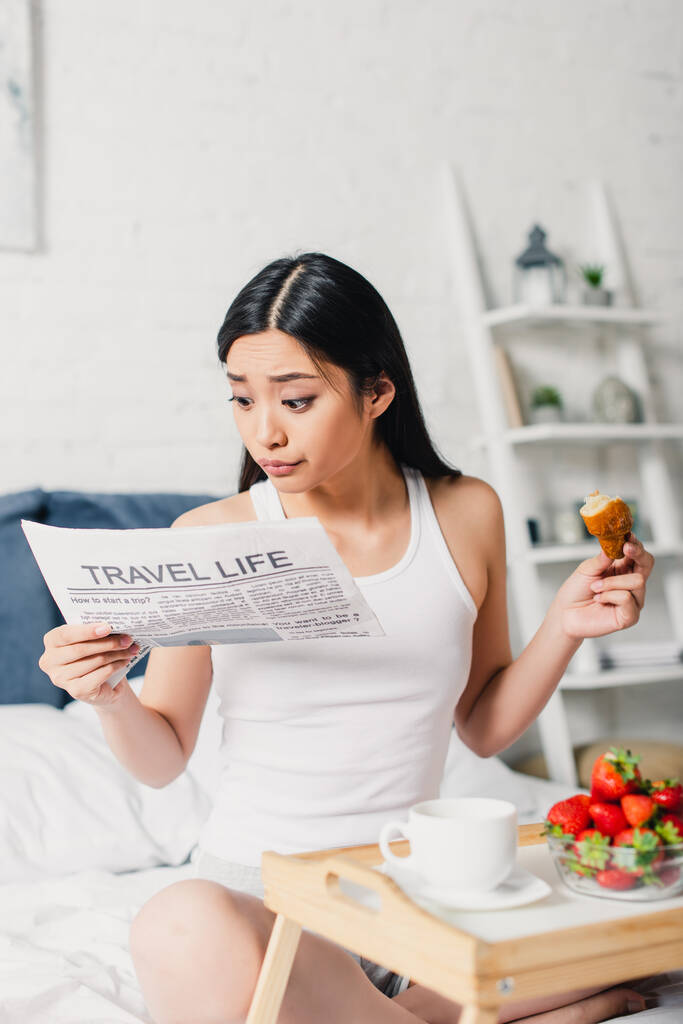 Selective focus of asian woman reading news and holding croissant near strawberries and coffee on breakfast tray on bed  - Photo, Image