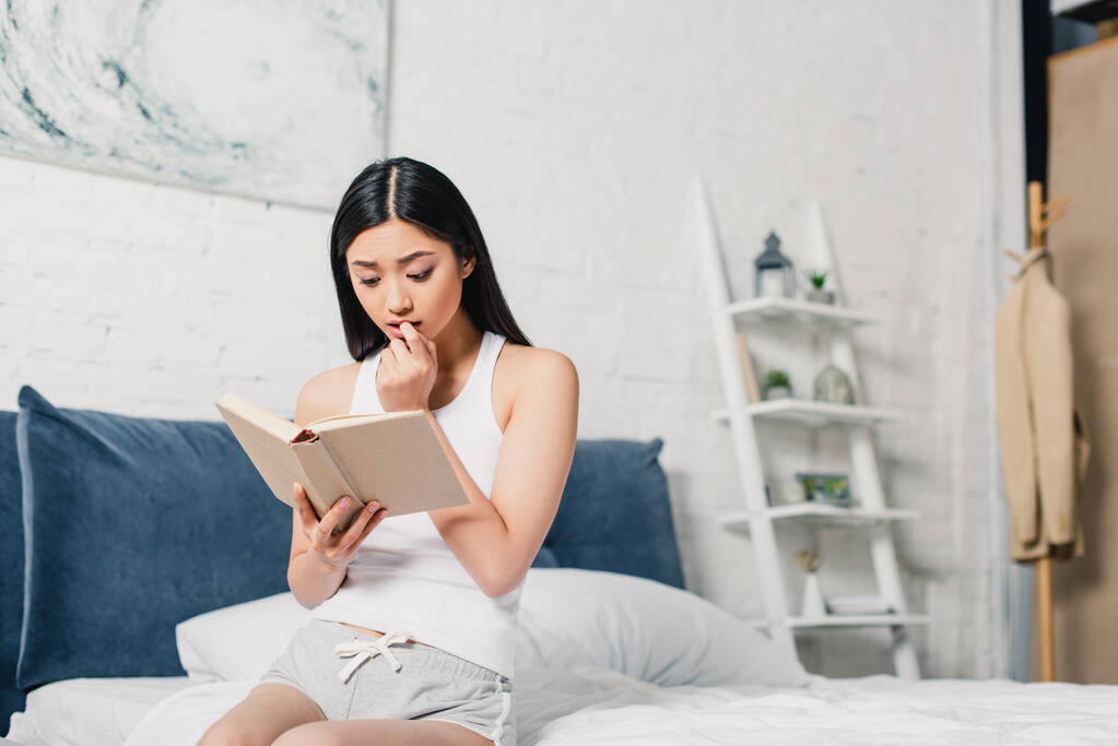 Concentrado asiático mujer lectura libro mientras sentado en cama  - Foto, Imagen