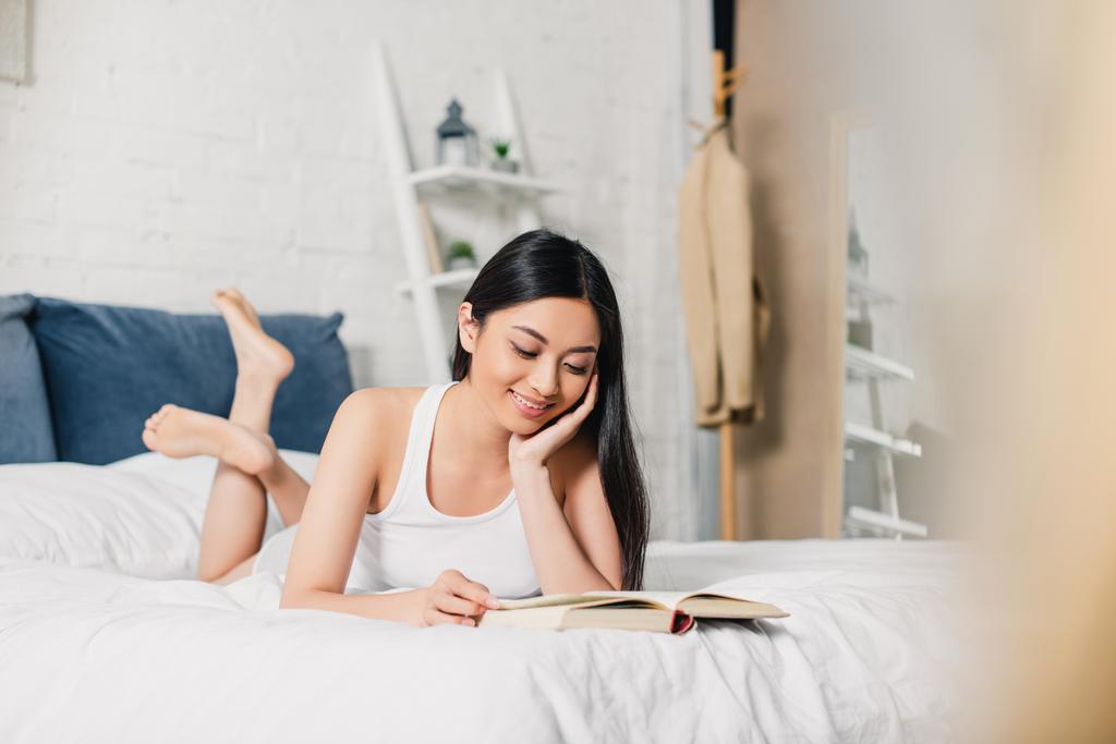 選択的焦点の笑顔アジアの女の子読書中にベッドに横たわっホーム  - 写真・画像