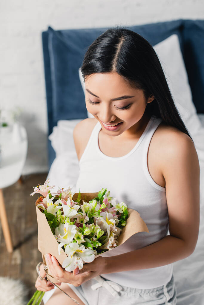 Smiling asian woman holding bouquet while sitting on bed  - Photo, Image