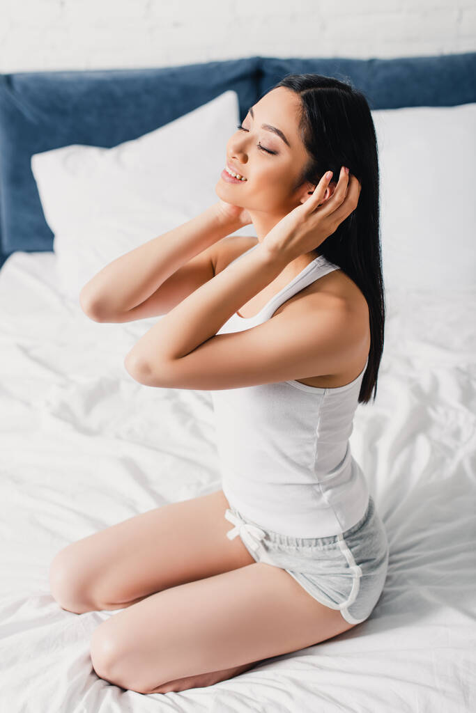 Sorridente ragazza asiatica toccare i capelli mentre seduto sul letto  - Foto, immagini