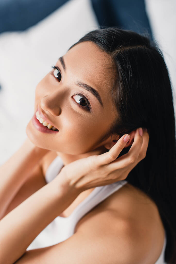 Vista aerea della giovane donna asiatica che sorride alla macchina fotografica mentre tocca i capelli sul letto  - Foto, immagini