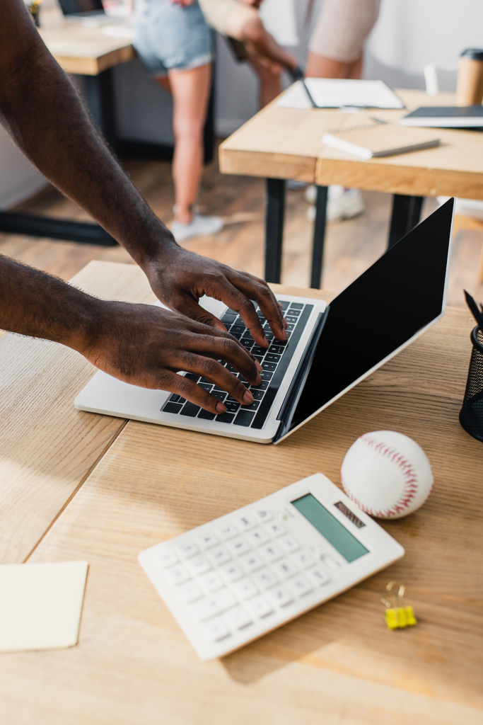 Vista recortada del hombre de negocios afroamericano usando el ordenador portátil con la pantalla en blanco cerca de la calculadora y bola de béisbol en la mesa de la oficina  - Foto, imagen
