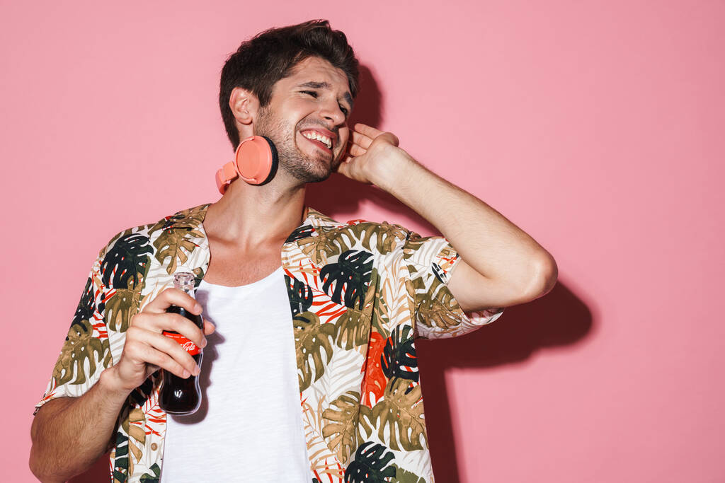 Portrait of cheerful young man using wireless headphones while drinking soda isolated over pink background - Photo, Image