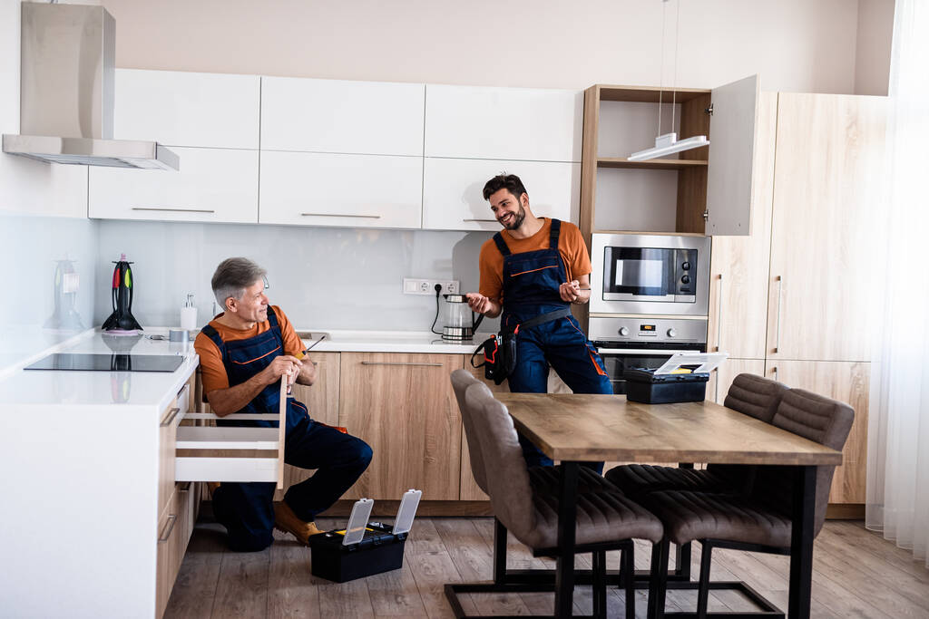 Tiro de longitud completa de dos manitas, trabajadores en uniforme hablando mientras ensamblan el gabinete de la cocina usando un destornillador en el interior. concepto de reparación y montaje de muebles
 - Foto, Imagen