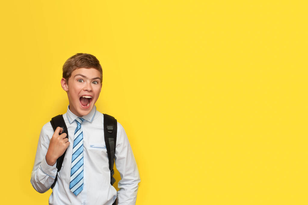 Um menino de camisa e gravata com uma mochila escolar emocionalmente se alegra, sorrindo em um fundo amarelo
 - Foto, Imagem