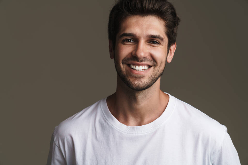Портрет радостного молодого человека в обычной футболке, улыбающегося и смотрящего на камеру, изолированную на сером фоне - Фото, изображение