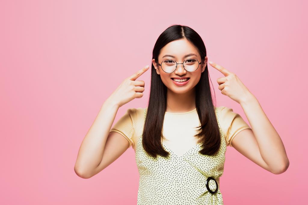 Ευτυχισμένη Ασιάτισσα που δείχνει με τα δάχτυλα τα γυαλιά που είναι απομονωμένα στο ροζ  - Φωτογραφία, εικόνα