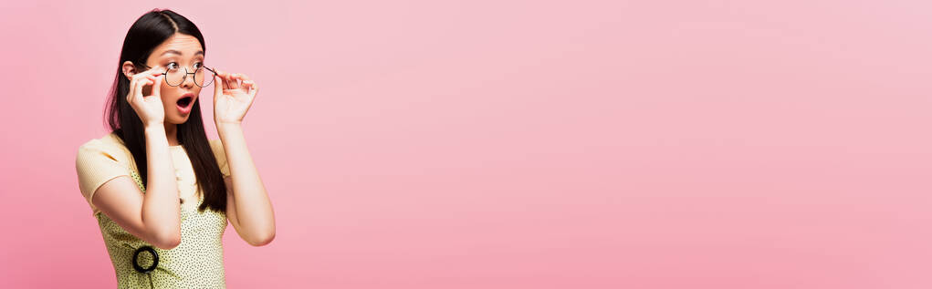 colpo panoramico di scioccato ragazza asiatica guardando lontano e toccando gli occhiali isolati su rosa  - Foto, immagini